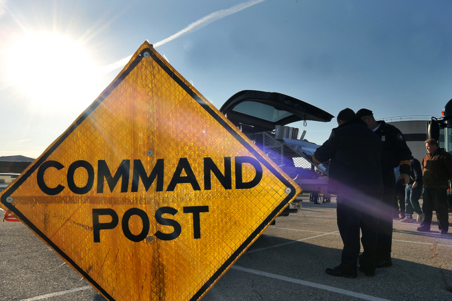 Orange temporary diamond road sign with "Command Post" on it, situated in a parking lot in front of a vehicle with two people standing by the open tailgate, working on something that cannot be seen in the picture.