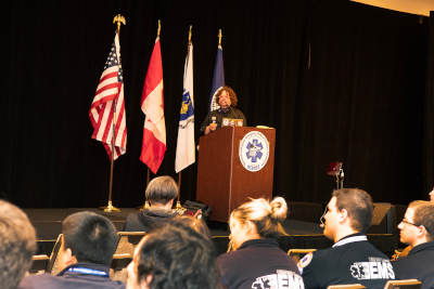 Dr. Ben Abo giving a keynote lecture, standing behind a podium with the NCEMSF logo, and in front of US, Canadian, Massachusetts, and NCEMSF flags. 
