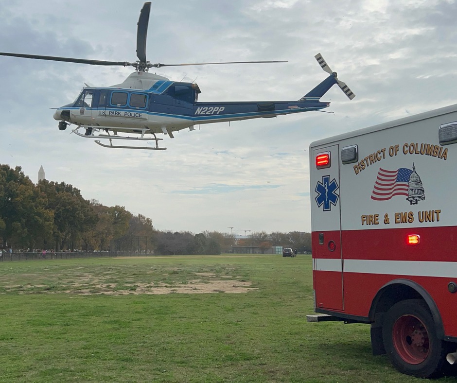 A US Park Police helicopter (blue, with white belly and main rotor engine compartment) hovering over a grassy field in a park. In the background are trees. To the right in the foreground is a white ambulance with two lower red stripes, the text District of Columbia Fire and EMS Unit above and below a graphic of a US flag, and the blue star of life emblem.