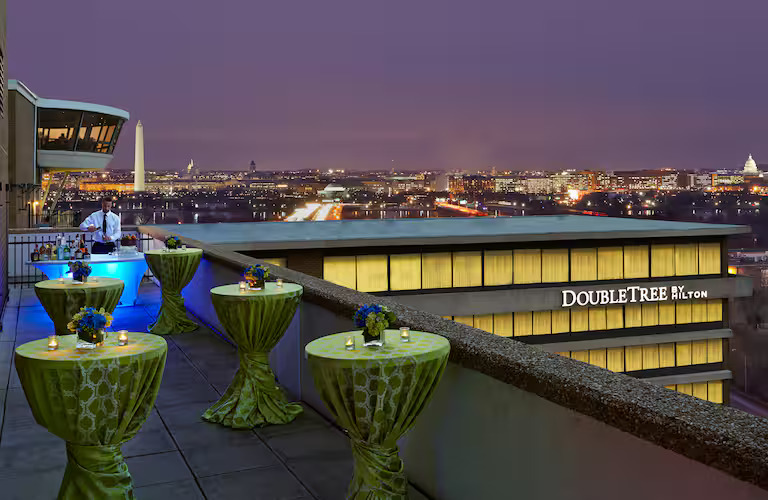 A view from the hotel's Jefferson Balcony at night. In the foreground are several round cocktail tables skirted in green patterned tablecloths, each with two small candles in glass jars and a small blue floral arrangement. At the far end of the balcony, a man in a white shirt mixes a drink while standing over a rectangular table skirted in white cloth with multiple liquor bottles arranged neatly on the left end. Above the man's head, you can see a portion of the circular rotating restaurant that sits atop the building. To the right, the top of the other wing of the hotel is visible, with the words DoubleTree by Hilton illuminated in white. In the background, you can see portions of Arlington and Washington, DC, including the Washington monument just to the right of the hotel restaurant, and the US Capitol dome can be seen in the far right of the photograph's background.