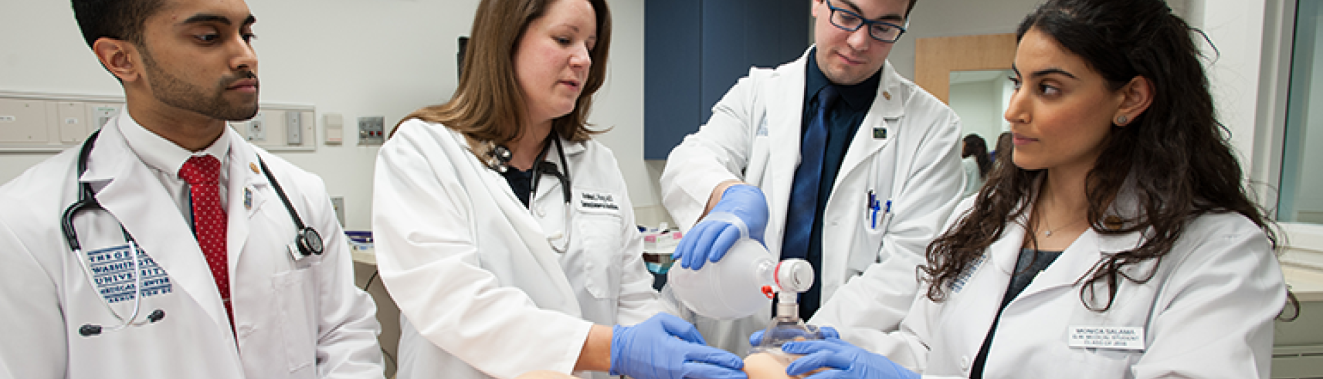 Four students in white coats stand around a patient. The student on the left watches with a stethoscope around his neck as the other three hold a bag-valve-mask device to a simulated patient's face and provide positive-pressure ventilation.