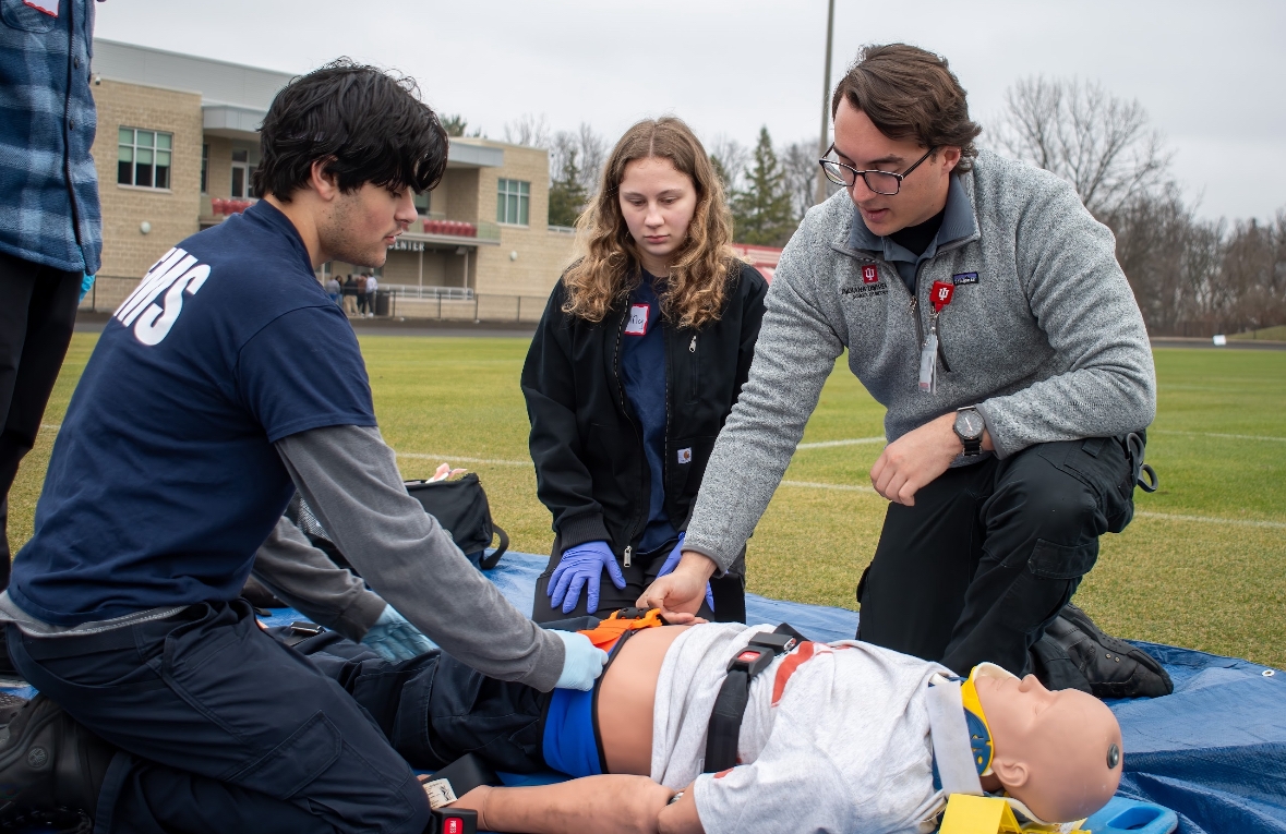 Three people kneel on the ground n a grassy field tending to a medical mannequin that is secured to a backboard.