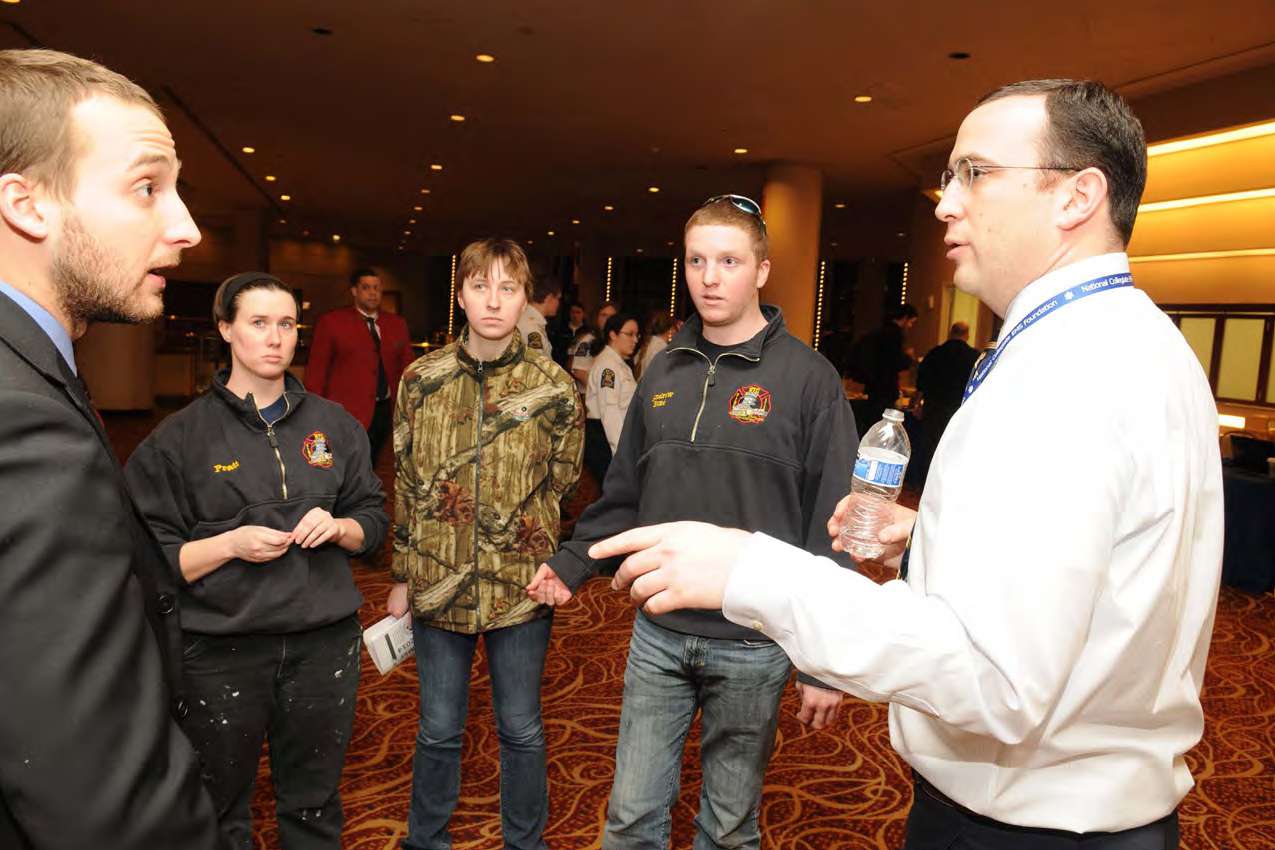 A man in a white collared shirt wearing a lanyard and holding a water bottle answers questions from a group of four other individuals. 