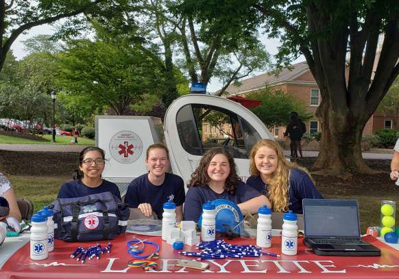 Four individuals sit at a table with a red tablecloth that says Lafayette on it. The table has a variety of objects with EMS branding, including water bottles and a first aid bag. Behind them is  a small electric vehicle with the EMS agency logo on the rear cargo box. 