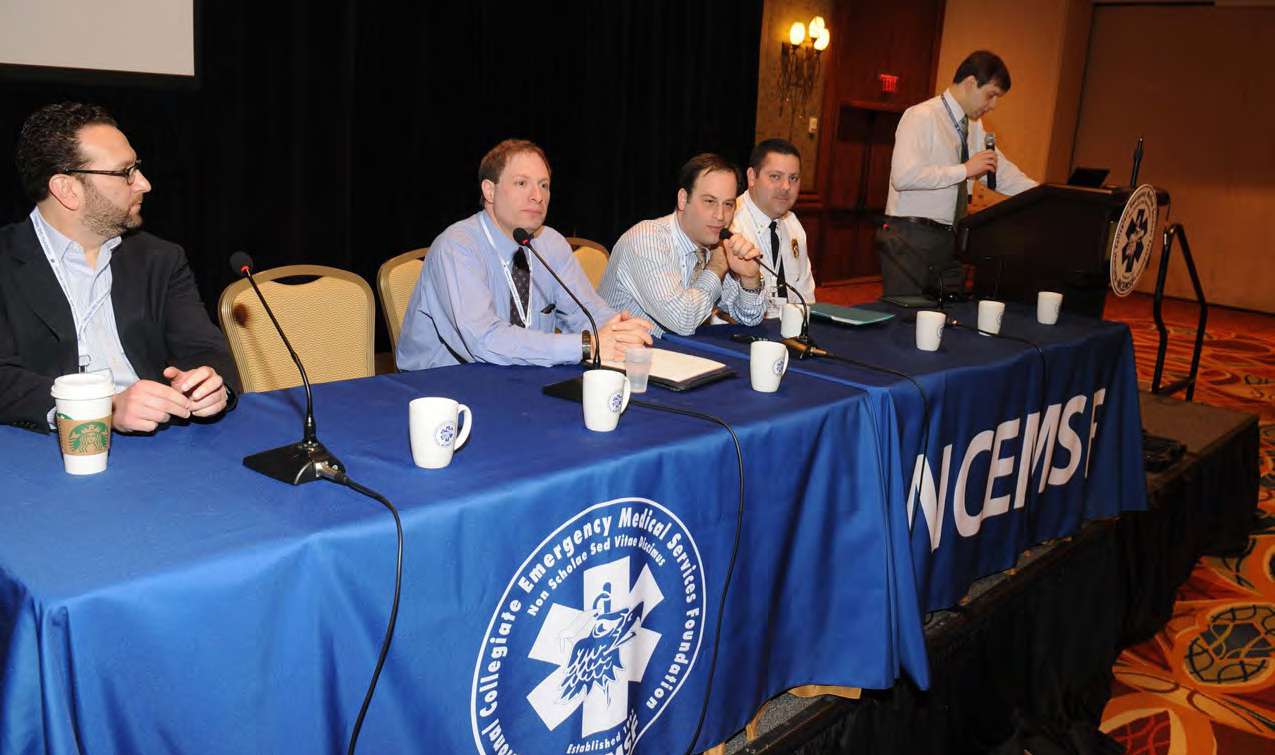 A group of five men participate in a panel discussion on stage at a conference. Four of the men are seated behind a row of tables covered in blue tablecloths with the NCEMSF logo and banding. Microphones and coffee cups sit atop the table. On the far right, the fifth man stands behind a lectern. 