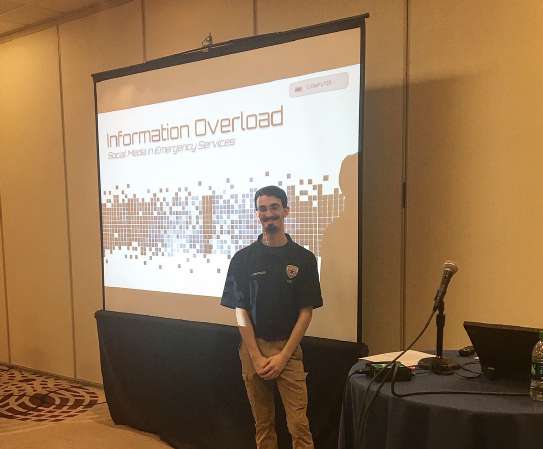 A person in a uniform polo shirt stands in front of a screen in a conference lecture room. Shown on the screen is the title slide of a presentation entitled Information Overload: Social Media in Emergency Services.