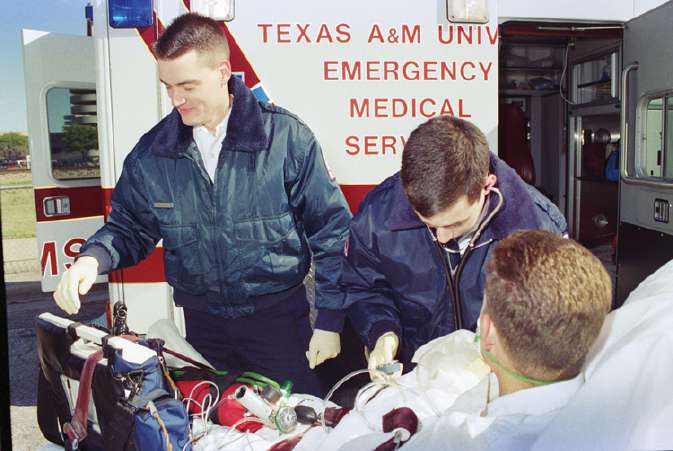 Two individuals wearing blue jackets tending to a patient on an ambulance cot. The cot has a variety of tubes and wires on it. Behind them is an ambulance with Texas A&M University Emergency Medical Services written on it side.