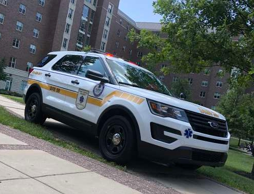 A white EMS quick-response SUV is parked on the grass in front of a university building. The SUV has red and white lights on the top and a yellow stripe down its side. 